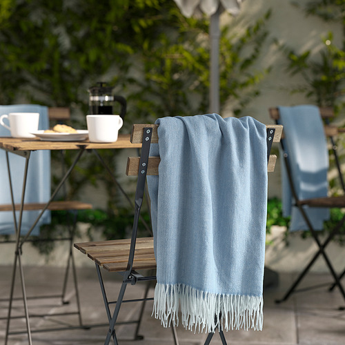 Manta HOLMVI azul claro con flecos blancos colgada sobre una silla, cerca de una mesa de madera con configuración de café, en un patio al aire libre.