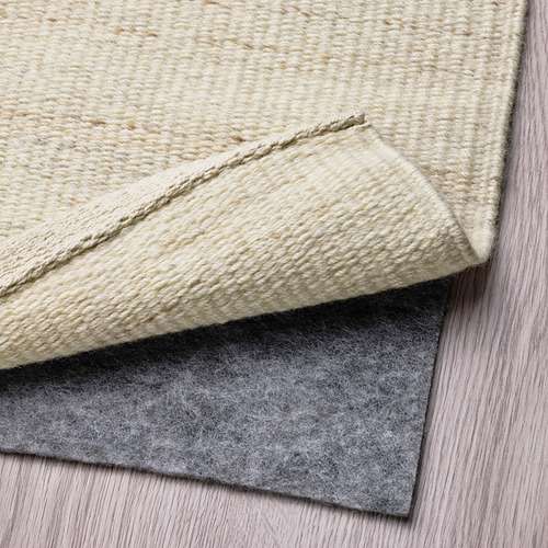 Un primer plano de una alfombra de tejido plano beige y su base antideslizante gris, que muestra la textura de la alfombra y cómo la capa de base la fija al suelo.