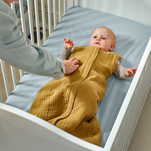 Un bebé en un saco de dormir amarillo yace en una cuna, con una mano de adulto cerca.