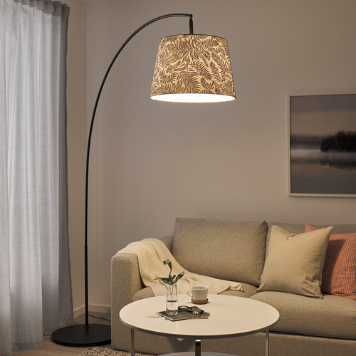 La lámpara de pie SKAFTET negra con pantalla de estampado de cebra cuelga sobre el sofá beige y la mesa blanca. La iluminación suave y cálida ilumina el acogedor rincón de la sala de estar.