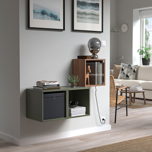 Mueble de pared EKET verde oscuro con 3 compartimentos, puerta de cristal y adorno de esfera superior. Debajo dos cuadros ondulados, cerca de un gran ventanal.