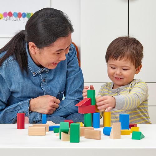 Un niño pequeño y un adulto juegan con bloques de madera de colores sobre una mesa blanca. El niño los está apilando, aprendiendo formas y colores.