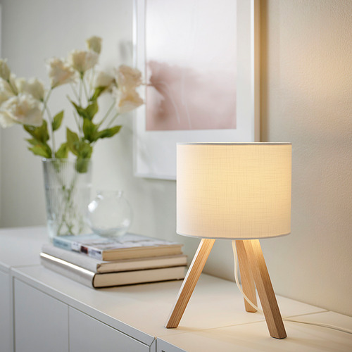Pequeña lámpara STORSEGEL blanca con base de trípode de madera, junto a flores y libros, luz cálida.