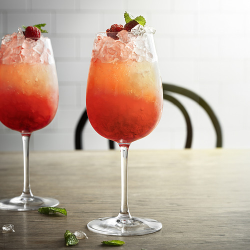 Dos copas altas de vino llenas de naranja, líquido rojizo, hielo, frambuesas y hojas de menta, colocadas sobre una mesa de madera.