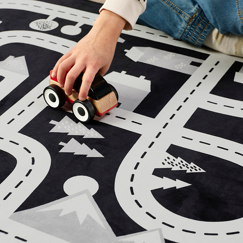 Coche de madera con dirección manual para niños en alfombra de juego en blanco y negro con diseños de ciudades y bosques.