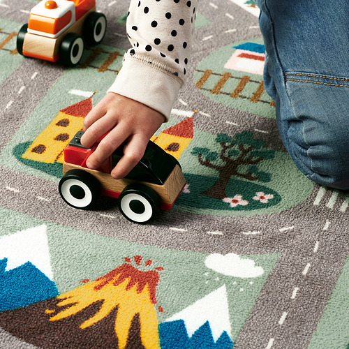 La mano del niño juega con el coche de madera sobre una alfombra colorida, mostrando su característica antideslizante.