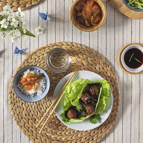 Una configuración de comida con un plato con envolturas de lechuga, un tazón de arroz, palillos y platos en un mantel individual tejido.