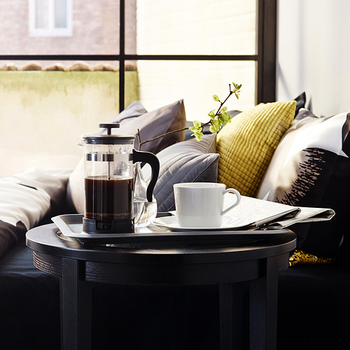 Prensa de café moderna en mesa negra con taza, que ofrece café recién hecho, ubicado cerca de un acogedor banco acolchado.