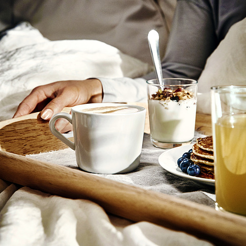 Sostiene de mano una taza de café espumoso en una bandeja con yogur, jugo y panqueques.