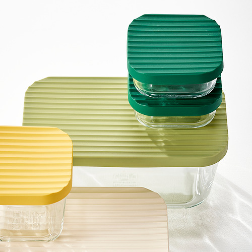 Tres coloridos recipientes de vidrio apilables con tapas texturizadas. Uno amarillo, otro verde y otro transparente, dispuestos sobre una superficie de madera.