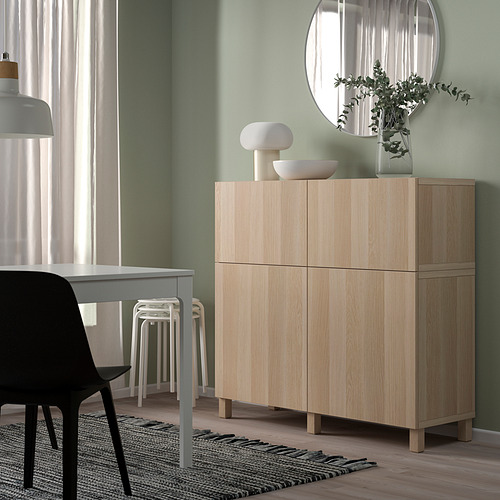Mueble BESTÅ de madera clara contra pared verde, con espejo redondo, silla negra y taburetes blancos.