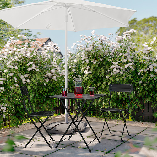 Patio plegable negro con mesa y sillas bajo una sombrilla blanca, ubicado en un patio de baldosas, rodeado de arbustos en flor.