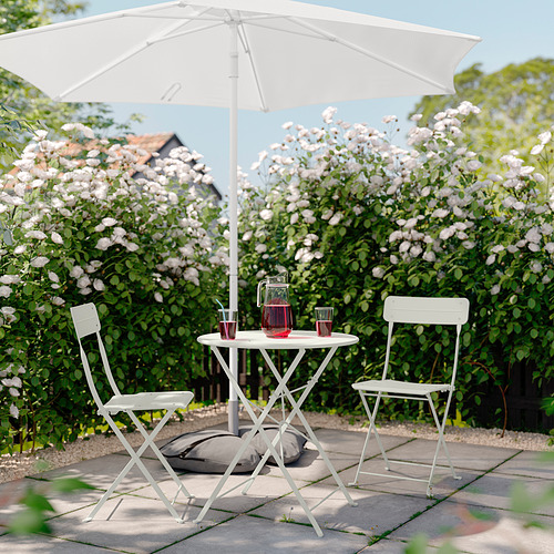 Serena escena de jardín con mesa y sillas blancas de SUNDSÖ, adornadas con bebidas, sobre un fondo de flores en flor.