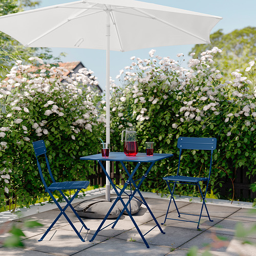 Patio de metal azul con mesa y sillas bajo una sombrilla blanca, ubicado en un exuberante jardín.