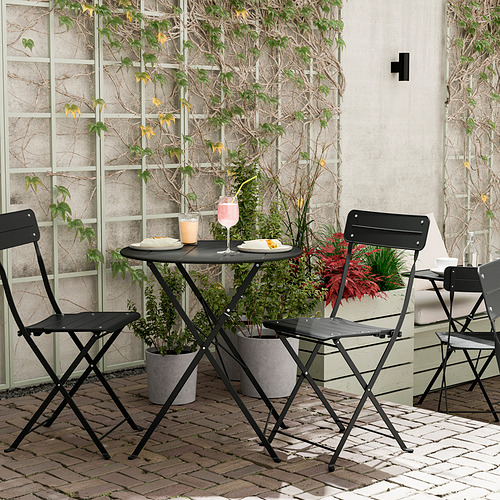 Conjunto de balcón de metal negro con mesa y sillas plegables, plantas en macetas.