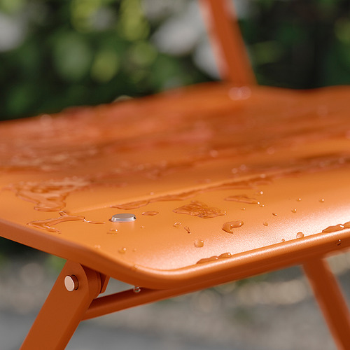 Silla plegable SUNDSÖ naranja en primer plano. La superficie brillante y húmeda con gotas de agua refleja la luz. Construcción duradera de acero con recubrimiento en polvo.