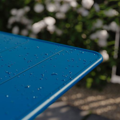 Mesa de exterior azul con gotas de agua, parcialmente plegada, sobre la vegetación.