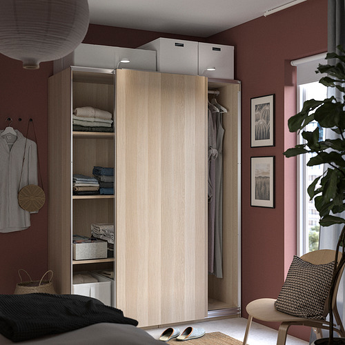 Dormitorio con armario PAX de madera de color claro, abierto para mostrar ropa y prendas dobladas, junto a una ventana con una planta y una silla.
