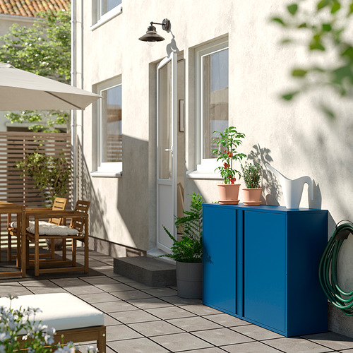 Pequeño balcón con gabinete de almacenamiento verde azulado, plantas en macetas, mesa de madera y asientos blancos al aire libre debajo de un edificio beige.