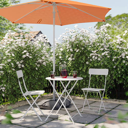 Pequeña configuración al aire libre con sombrilla naranja, mesa blanca y dos sillas rodeadas de arbustos en flor.