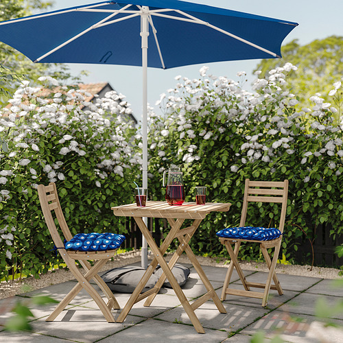 Relajado patio al aire libre con cojines de lunares azules en sillas de madera, sombrilla azul, flores blancas, bebidas en mesa de madera.
