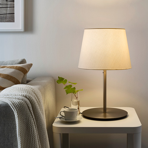 Una lámpara SKOTTORP con pantalla beige en la mesa auxiliar, rodeada de taza, hojas de parra y un acogedor sofá con manta blanca. Pantalla de plástico suave y brillante.