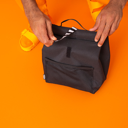 Persona sosteniendo una pequeña bolsa de almuerzo negra con rayas blancas sobre fondo naranja.