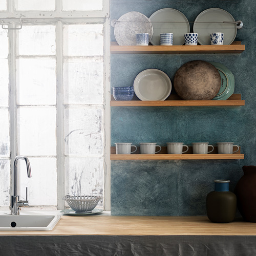 Encimera de cocina con fregadero blanco, tazas y jarrones azules, platos y un cuenco en estantes de madera contra la pared azul.