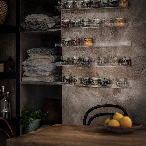 Los estantes flotantes de acrílico transparente sostienen frascos de vidrio con alimentos en conserva, pared metálica, mesa de madera con limones y silla.