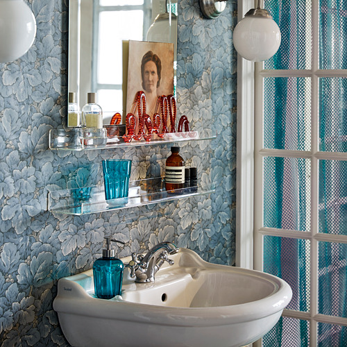 Baño con papel pintado floral azul, lavabo blanco, estantes de vidrio transparente con varios artículos de tocador y una foto enmarcada.