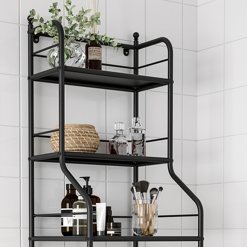 Estantería de baño de metal negro con tres niveles que almacenan toallas, botellas, jarrones y brochas de maquillaje contra la pared de azulejos blancos.