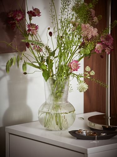 Un jarrón PÅDRAG de vidrio transparente con un diseño redondo y acanalado, que sostiene un ramo de flores rosas y blancas con tallos y hojas verdes.