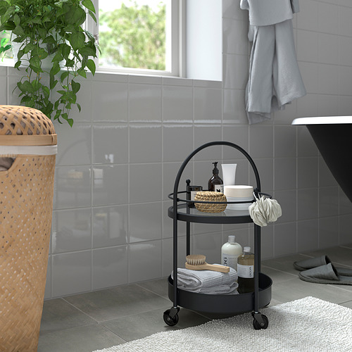 Pequeño carrito de baño FRÖSJÖN de metal negro con tapa de cristal, con toallas, cepillos y cremas, junto a una gran cesta de mimbre y azulejos grises.