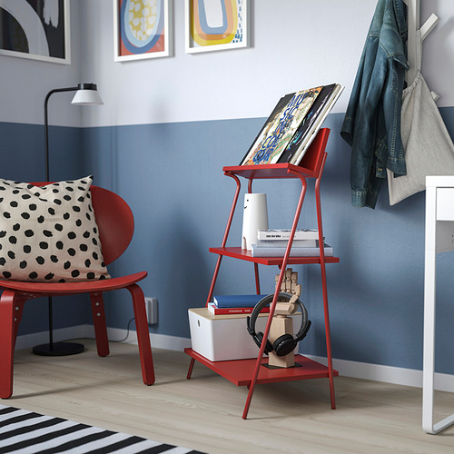 Estantería roja KRÖNIKÖR con libros, altavoz y silla con almohada contra la pared azul, perchero cerca.
