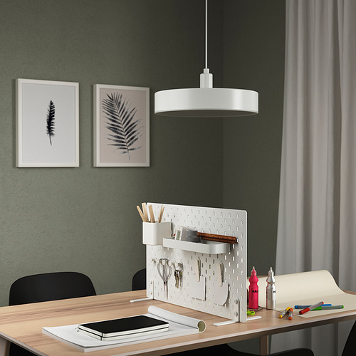 El separador de escritorio blanco SKÅDIS se mantiene alto, organizado con suministros de oficina, arte enmarcado en la pared, iluminado por una lámpara colgante.