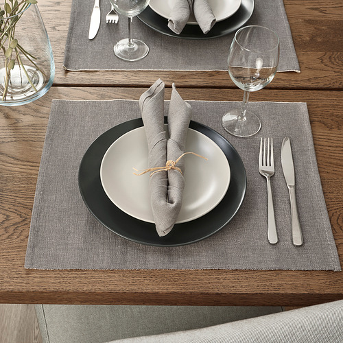 Mesa con manteles individuales grises SVARTSENAP, platos con borde negro, servilletas dobladas, cubiertos y copa de vino.