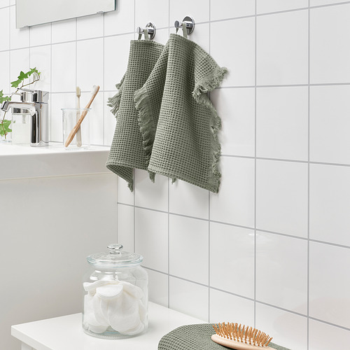 Las toallas verdes VALLASÅN cuelgan de la pared del baño con azulejos blancos, con un mostrador que contiene bastoncillos de algodón, cepillos de dientes y una planta.