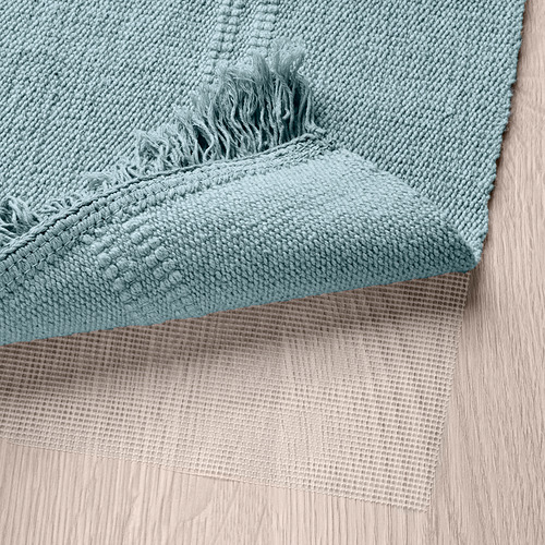 Alfombra de algodón tejido verde, reversible, lavable a máquina, colocada sobre suelo de madera. Detalle de flecos a la vista.