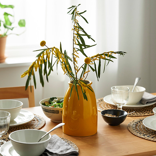 Una mesa de madera con cuencos blancos, cuencos blancos y negros, un vaso y un jarrón amarillo con ramas verdes y flores amarillas.