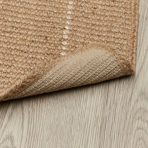 Esquina de alfombra de yute beige sobre suelo de madera, con reverso antideslizante texturizado.