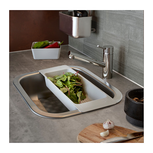 Fregadero de cocina moderno con grifo de acero inoxidable. Colador blanco con verduras verdes en el fregadero. Tabla de cortar de madera con cuchillo y ajo cerca.