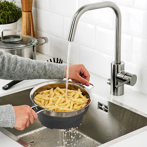 Persona enjuagando pasta cocida en un fregadero de acero inoxidable con un colador de silicona flexible con agua corriente.