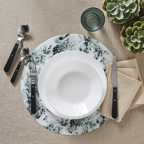 Elegante mesa con cuenco blanco, plato floral, utensilios con mango negro, servilleta beige y cristal verde sobre mantel individual beige texturizado.