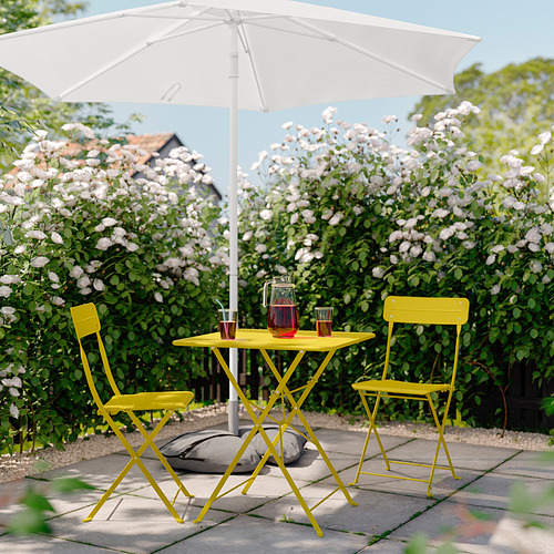 Mesa y sillas de exterior de color amarillo brillante con sombrilla blanca, rodeadas de arbustos en flor, ideales para espacios pequeños.
