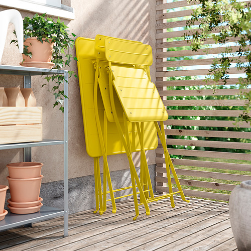 Sillas plegables de metal SUNDSÖ de color amarillo brillante, apiladas ordenadamente al aire libre contra una pared. Perfectos para espacios pequeños con su diseño duradero y fácil de plegar.