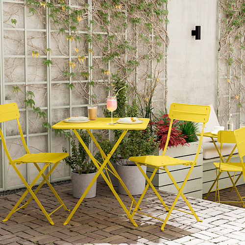 Conjunto de patio plegable de color amarillo brillante con una pequeña mesa y sillas sobre suelo de ladrillo, rodeado de plantas en macetas y una pared beige con una celosía.