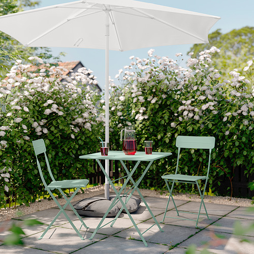 Escena de patio sereno con mesa y sillas de metal SUNDSÖ verde claro, sombrilla blanca, flores rosas y bebidas.