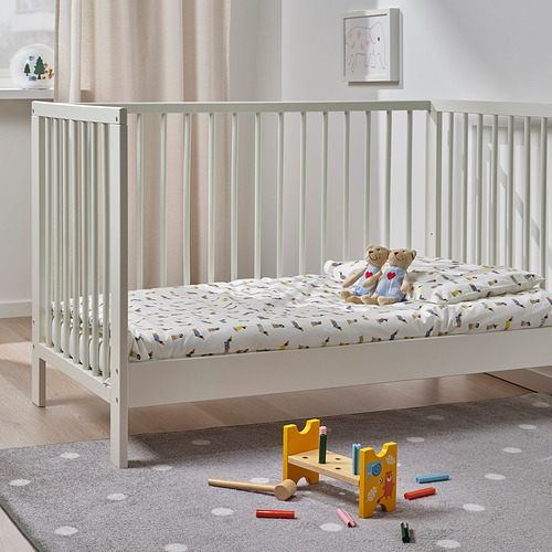 Cama infantil juguetona con estampados y peluches, sobre una alfombra gris, cerca de un juego de martillos de juguete.