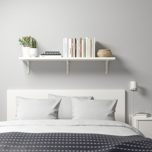 Dormitorio moderno y minimalista con estante de pared blanco que muestra libros, plantas y decoración sobre una cama con almohadas blancas y manta gris.