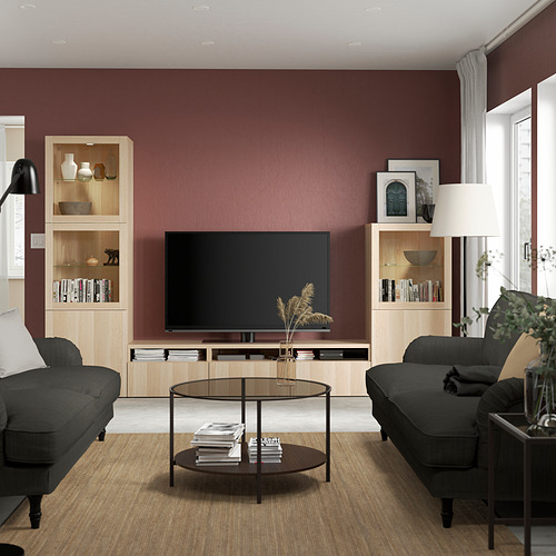 Sala de estar moderna con paredes granates, TV de pantalla plana en soporte BESTÅ, dos sofás grises, mesa de centro de vidrio y lámpara de pie alta.